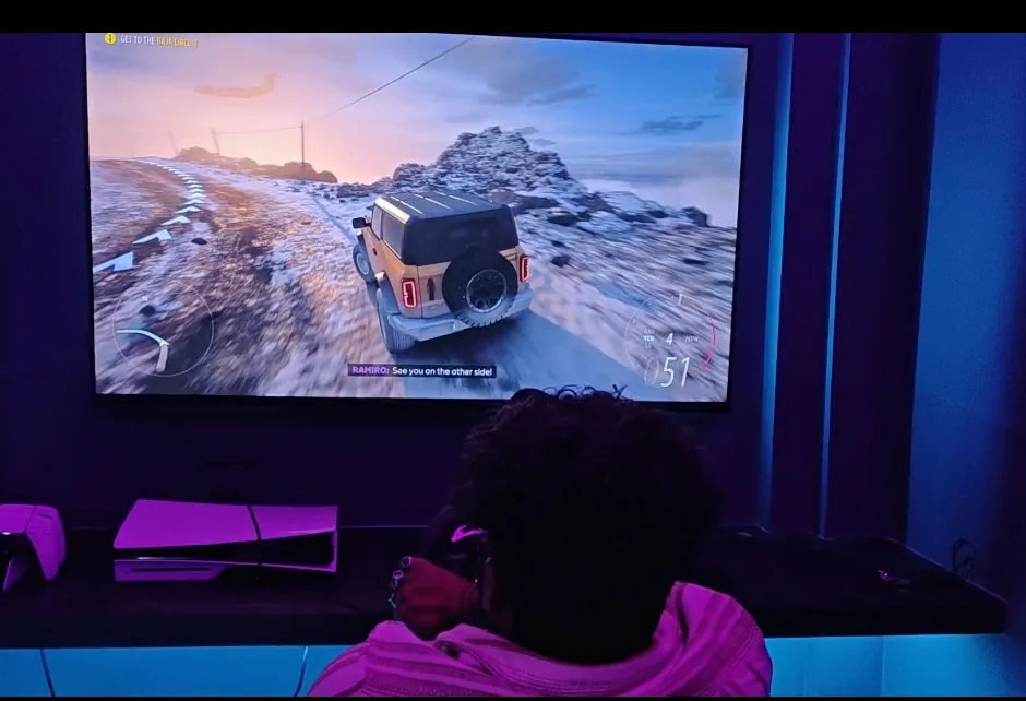 A player driving through snowy off-road terrain in a racing game on a large display with a PS5 below